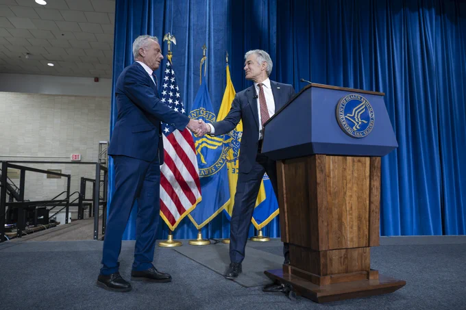 Administrator Oz shakes Secretary Kennedy's hand while standing at a podium. 