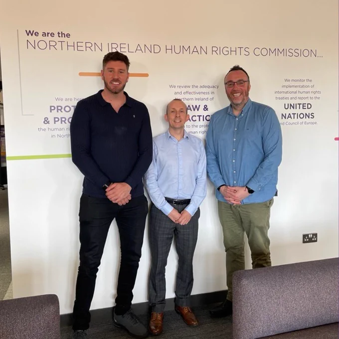 Three men are standing in front of a wall that reads "We are the Northern Ireland Human Rights Commission"