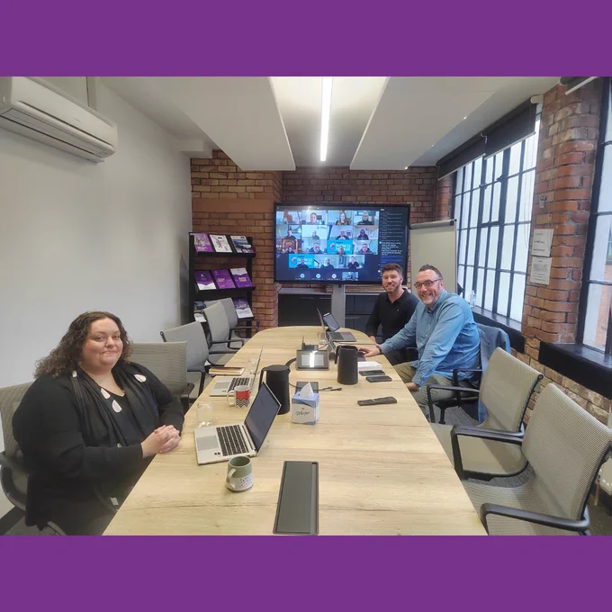 Photo of a boardroom with three people sitting in person, in the background a screen with multiple people online can be seen.