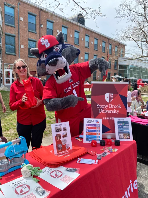 Stony Brook University mascot in red outfit poses with a person at an outdoor library information booth on campus lawn.