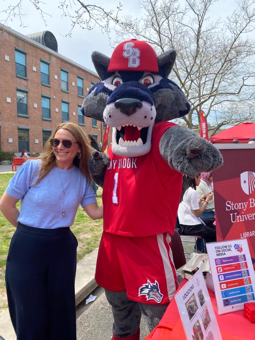 Stony Brook University mascot in red jersey posing with a woman outdoors near campus event booths and a university banner.