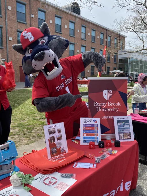 Stony Brook University mascot in red shirt and hat poses behind a table with university brochures and promotional items outdoors on campus.