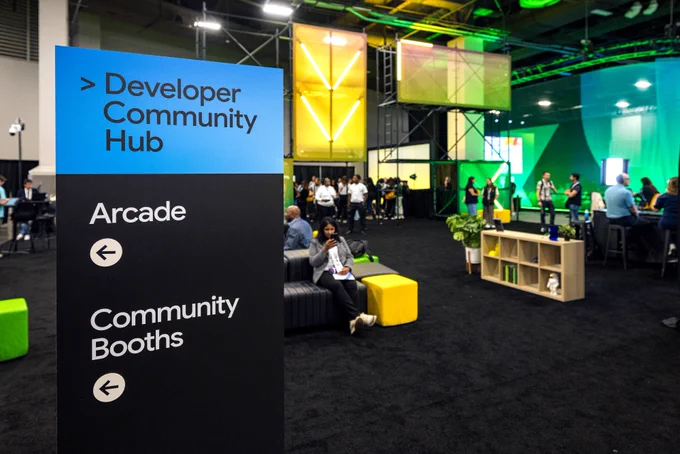 A large directional sign at a tech event prominently displays "Developer Community Hub" in a blue section, and "Arcade" and "Community Booths" with left-pointing arrows in a black section below. In the background, people walk through a modern lounge area with black carpeting, gray and yellow seating, and large, brightly lit yellow and green translucent partitions. One person with dark hair sits on a gray couch in the midground, wearing a gray blazer and looking at their phone.