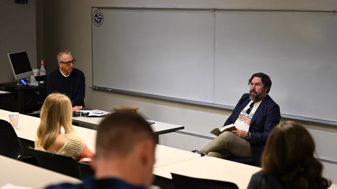A classroom scene with two individuals at the front. One is seated, holding a book, while the other sits nearby, engaged in conversation. Several people are seated in rows facing them. The setting is a lecture room with a large whiteboard.
