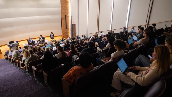 This image shows a large lecture hall at Shaddock Auditorium in the Neeley School of Business filled with attendees. The audience is seated, facing a stage where several individuals are involved in a discussion. The room has tiered seating and wooden accents, and many attendees have laptops open.