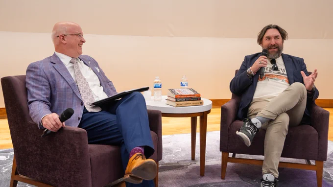 Two individuals are seated on purple chairs, engaging in a conversation. A stack of books and two water bottles are on a small table between them. One person is in a plaid suit and is smiling, while the other is speaking and gesturing, wearing casual attire.