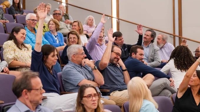 An audience in a lecture hall, with several attendees raising their hands. Some people appear engaged while others look towards the speaker. The setting is academic and formal.