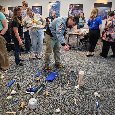 A group of adults participate in a hands-on workshop in a conference room. In the center, a man wearing a bomber jacket with space mission patches operates a simulated robot arm, pulling a string to guide the cardboard device across the carpeted floor. The floor is scattered with various objects. The other participants watch him, smiling. 