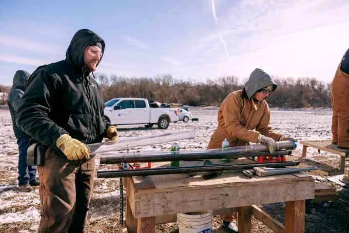 The image represents vadose coring. Two workers dressed in winter gear handle long metal rods on a wooden workbench outdoors with snow on the ground. 