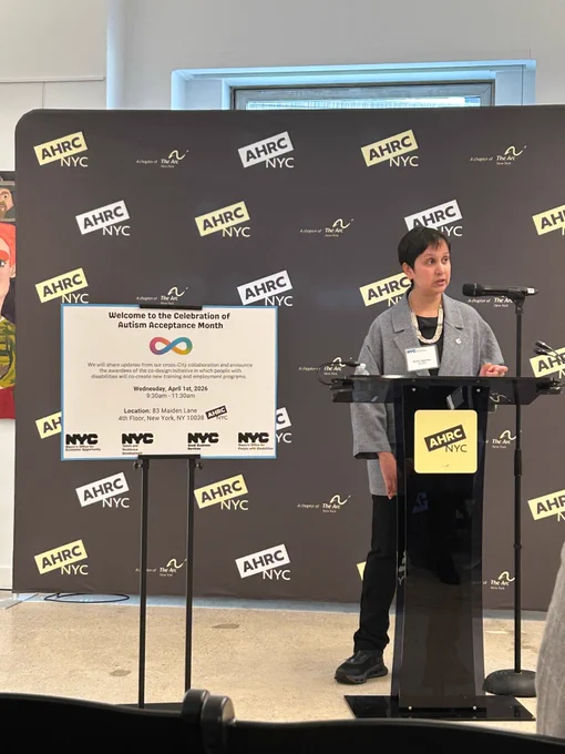 
Commissioner Agarwal is standing at a podium, speaking at a formal event. Behind her is a backdrop covered with the AHRC NYC logo. A sign on the podium welcomes attendees to a Celebration of Autism Acceptance Month. The setting feels official and community‑focused, highlighting advocacy for people with disabilities.