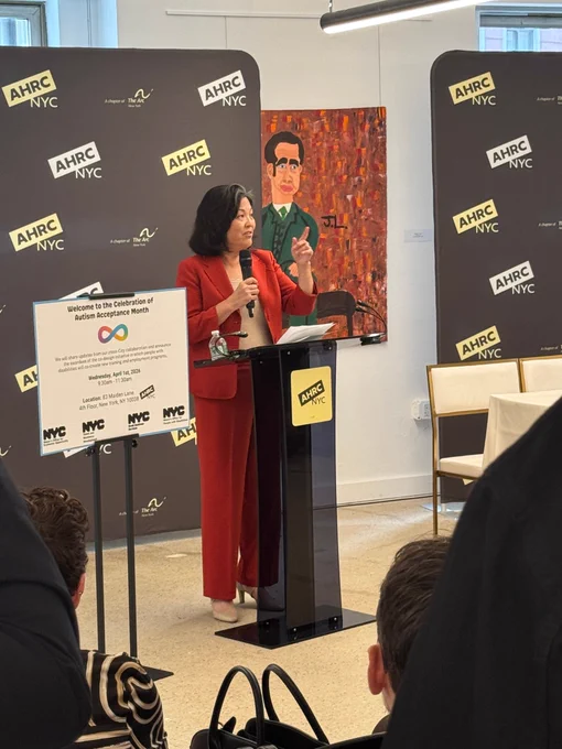 Deputy Mayor Julie Su is standing at a podium, speaking at a formal event. Behind him is a backdrop covered with the AHRC NYC logo. A sign on the podium welcomes attendees to a Celebration of Autism Acceptance Month. The setting feels official and community‑focused, highlighting advocacy for people with disabilities.