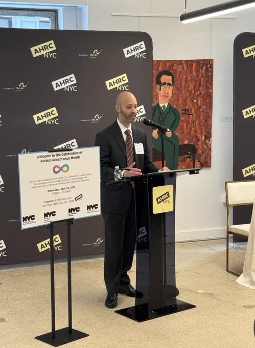 Commissioner Minaya is standing at a podium, speaking at a formal event. Behind him is a backdrop covered with the AHRC NYC logo. A sign on the podium welcomes attendees to a Celebration of Autism Acceptance Month. The setting feels official and community‑focused, highlighting advocacy for people with disabilities.