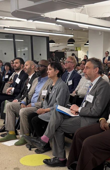 This image shows a group of people seated in rows, paying close attention to something happening off-camera. They’re dressed in business attire and wearing name tags, suggesting a conference or seminar. The room has a modern office look with glass partitions and bright lighting. It captures a moment of engagement and collaboration.