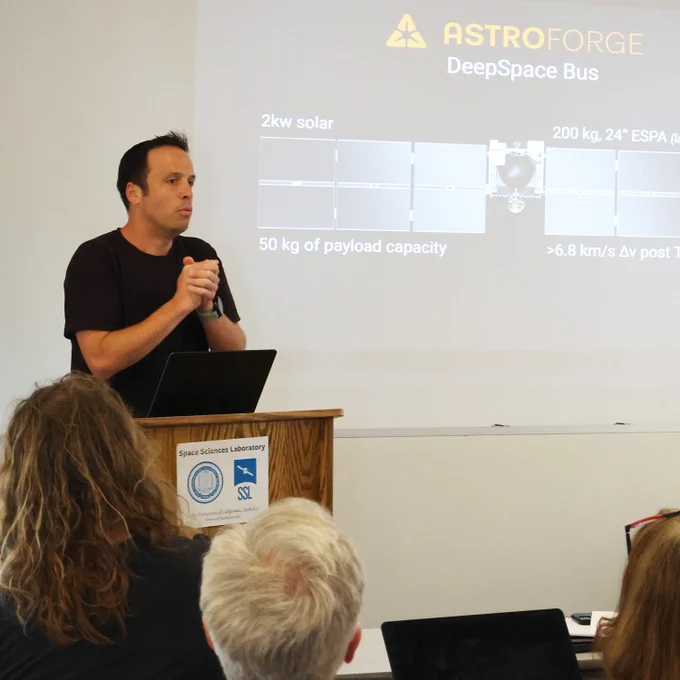 Matt Gialich, Founder and CEO of AstroForge, speaking behind the podium at the UC Berkeley Space Sciences Laboratory conference room and standing beside a slide displaying the AstroForge DeepSpace bus.