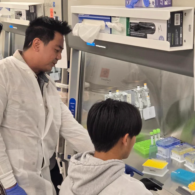 In a laboratory setting, individuals wearing white lab coats work at a lab bench surrounded by scientific equipment. Supplies from Thermo Fisher Scientific are visible.