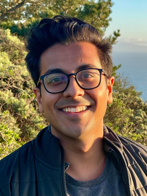 Headshot of Manu Meel '20 outdoors, with greenery and a skyline visible in the background. The sun is setting and illuminating the left side of his face.