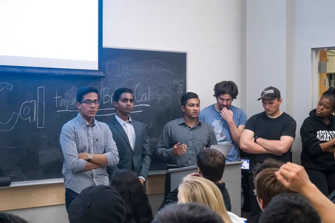 The early days of BridgeUSA. Meel and some of his classmates are standing in front of a podium in a classroom giving a speech. Behind them are a chalkboard with a TinyURL link and a projector screen.