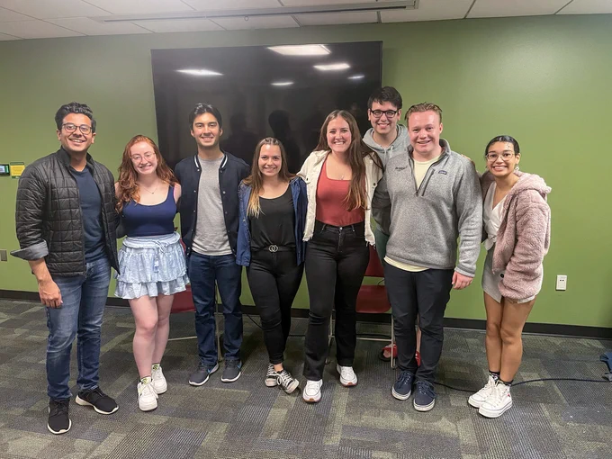 A group photo of Meel (left) and a handful of BrideUSA team members in an indoor environment. The wall behind them is a lime green color, and a tv is mounted on it.