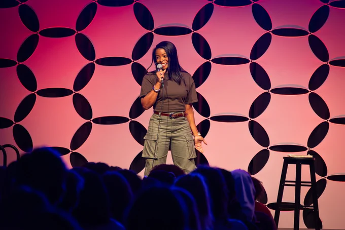 Comedian Janelle James performs on stage at RamFest, holding a microphone and smiling at the audience. She wears a brown top and olive cargo pants with a brown belt. Behind her, a geometric petal-patterned backdrop is lit in a pink-to-coral gradient. A stool with a notebook sits to her right. Silhouetted audience members fill the foreground.