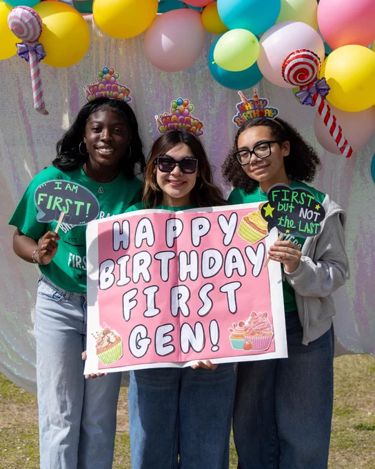 Three people are standing together holding a sign that reads "Happy Birthday First Gen!" They are wearing festive headbands and surrounded by colorful balloons.