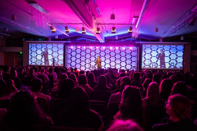 Wide shot of the Lory Student Center ballroom during Janelle James's RamFest performance. The packed audience faces the stage, which features the geometric backdrop flanked by two large projection screens displaying the performer. Pink and purple stage lighting washes over the room and crowd.