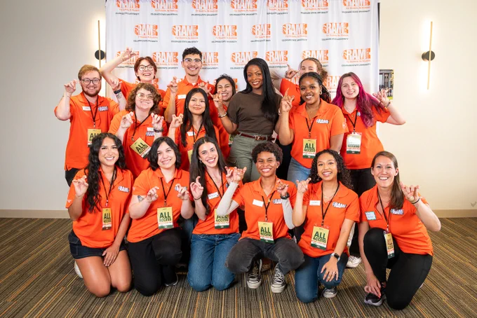 Larger group photo of the Ram Events student staff with Janelle James at the step-and-repeat. Approximately 17 people in orange Ram Events polo shirts pose in two rows, many throwing the "Rams up" hand sign. Janelle James stands in the back row center in her brown top and olive pants. Students wear "All Access" badges and lanyards.