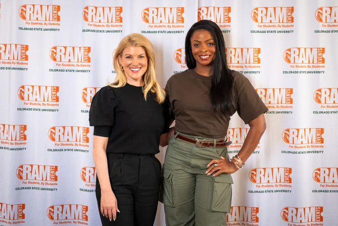 CSU President Amy Parsons and comedian Janelle James pose together in front of the Ram Events step-and-repeat banner. Parsons wears a black puff-sleeve top and black pants; James wears her brown top and olive cargo pants with silver bracelets. Both are smiling.