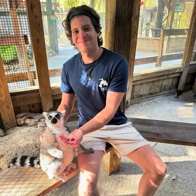 A man sitting down next to a ring-tailed lemur inside the ring-tailed lemur exhibit at Wild Florida Adventure Park.