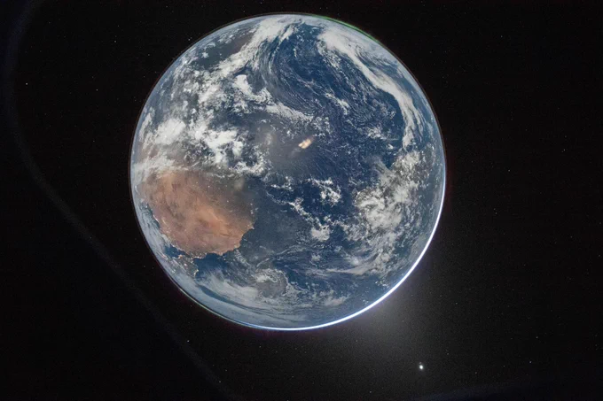 A full disk image of Earth, as seen from the Orion capsule. The planet is a pale blue, swirling with white clouds and glowing slightly lighter blue in place from reflected light. From about 8 to 9 o'clock, a large brown landmass is Africa, with the Iberian peninsula twinkling with lights just where the planet curves. At the 1 o'clock spot, aurora glow in a thin green glow, just barely separated from the planet's surface. Earth is set against the black of space. Credit: NASA/Reid Wiseman