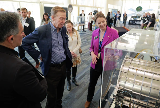 A group of people is gathered around a scientific display at a technology exhibition. LASP Director Bethany Ehlmann is explaining the exhibit, which includes machinery within a clear casing. There are various informational signs and banners in the background, including one that reads "LASP." The setting appears to be indoors with large windows allowing natural light.