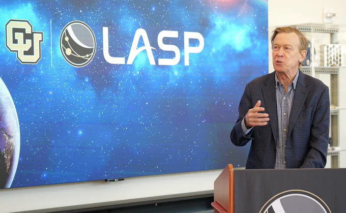 Senator John Hickenlooper stands at a podium giving a speech. Behind them, a large screen displays the University of Colorado logo and the LASP logo against a starry background.
