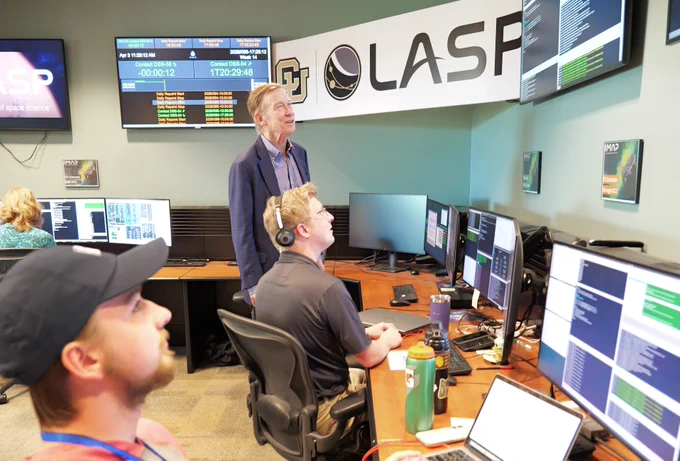 A group of individuals in a control room or office setting are focused on multiple computer monitors displaying various data and graphs. The room is part of LASP, as indicated by a prominent banner on the wall. Senator John Hickenlooper is standing and observing the screens while others sit at their desks, wearing headsets and working on the computers. The environment appears busy and technical.