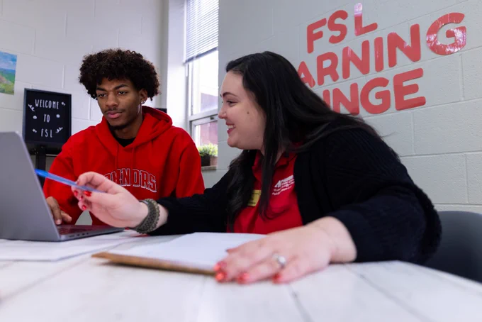Two people sitting at a table in the FSL Learning Lounge, working on a laptop. One person is holding a pen and pointing at the computer screen.