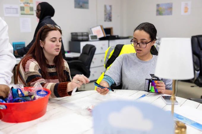 Two people are sitting at a table engaged in a creative activity, possibly organizing arts and crafts materials. The table is covered with various supplies, including markers and containers. In the background, there are more individuals and office furniture. The setting appears to be a community center or classroom.