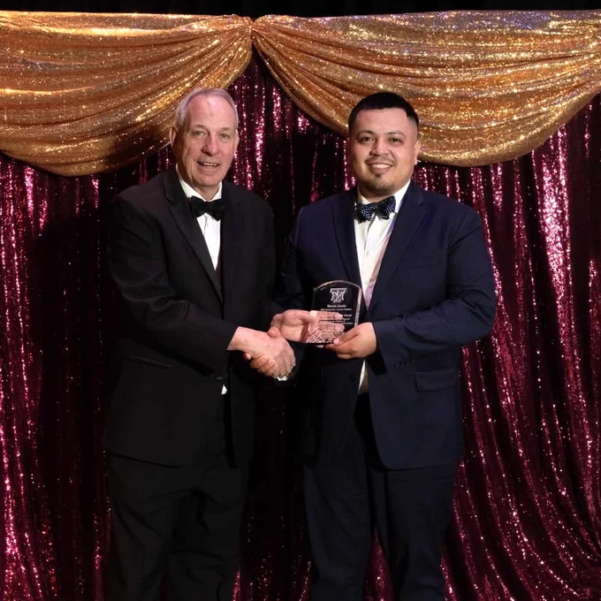 Two people in formal attire are shaking hands while a Texas A&M University-Corpus Christi alumnus is holding an award. They stand in front of a backdrop with shimmering gold and maroon drapes.
