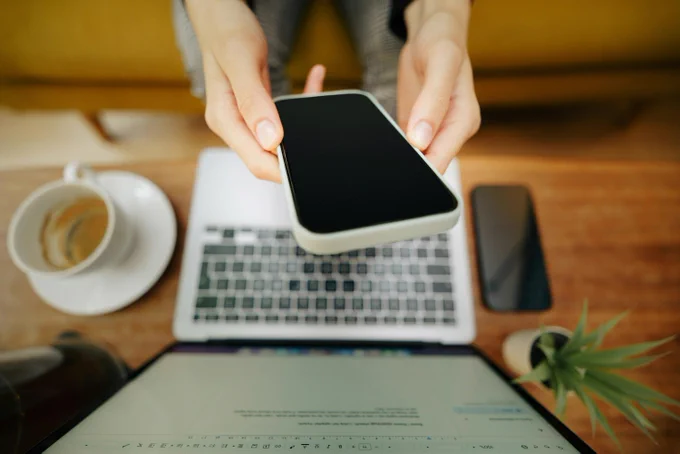 A close up of an individual using a laptop while holding a phone in front of it.