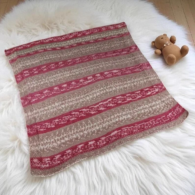 A knitted baby blanket lies on a fluffy white rug. The blanket has alternating horizontal stripes in shades of taupe and a reddish pink with subtle cream patterns within the coloured bands. The texture appears soft and cosy. To the right of the blanket, a small brown teddy bear with a ribbon around its neck rests on the rug.