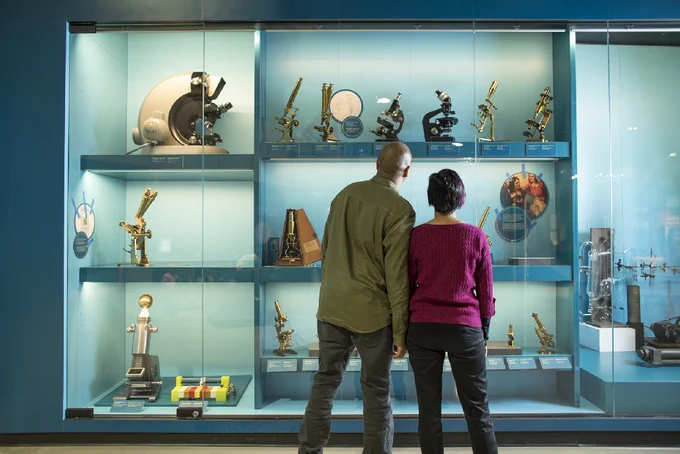 Deux personnes observent une vitrine de musée remplie de microscopes anciens et d'instruments scientifiques, sous un éclairage tamisé.