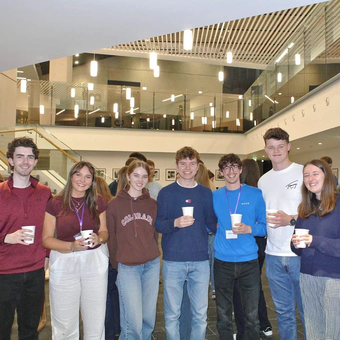Group of students standing indoors holding cups, socialising in Ulster University with glass railings and bright lighting.