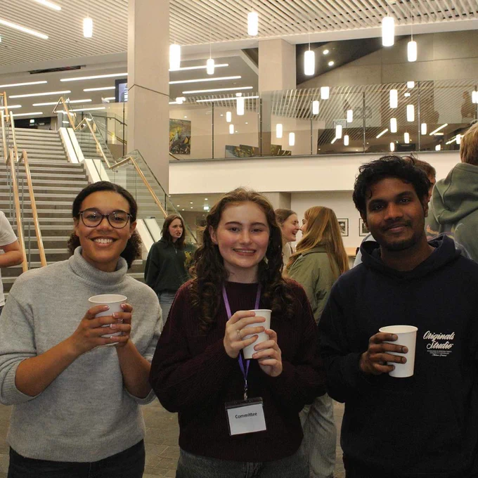 Three young students holding coffee cups in a modern building lobby with stairs and hanging lights in the background.