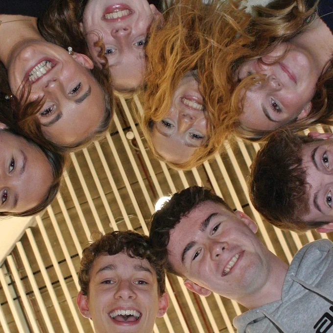 Group of students smiling and looking down at the camera in a circular formation under a wooden slatted ceiling.