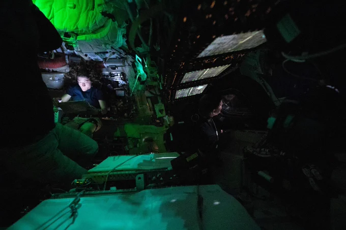 Astronaut Christina Koch is nestled into a corner of the Orion crew capsule, reading. The rest of the capsule is dark, but her face it lit by the glow from her tablet. Two of her other crew members can dimly be seen looking out the window and floating in the foreground. Credit: NASA