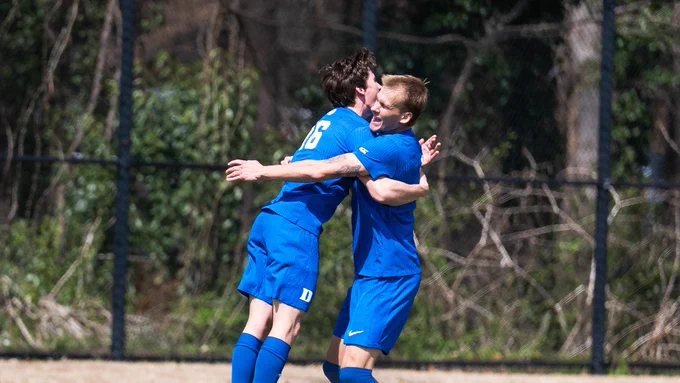 Two soccer players in blue jerseys embrace each other in celebration on a grassy field. They are smiling and showing signs of excitement after a successful play. The background features trees and a fence, indicating an outdoor sports environment.