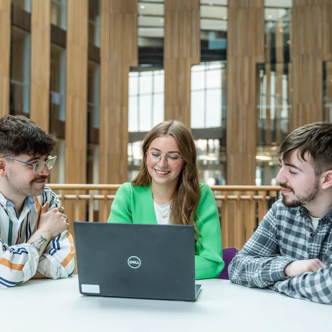 Three young adults sit around a table with a laptop, engaged in a discussion inside a modern building with wooden pillars and large windows.

