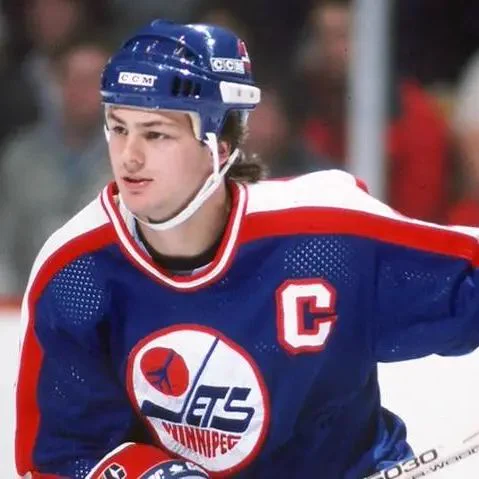 A color photo of Dale Hawerchuk skating on the ice during a game. He wears a blue Winnipeg Jets jersey with red and white accents, featuring the Jets logo on the chest and a captain’s C on the upper left. He also wears a blue helmet and gloves, holding a hockey stick as he looks ahead. The background shows a blurred crowd and rink boards.
