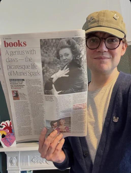 A photo of me, holding up a copy of The Times which features a review of my book, accompanied by the headline: ‘A genius with claws - the picaresque life of Muriel Spark’