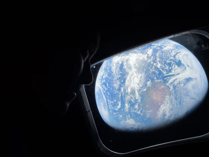 Inside the dim Orion spacecraft, a window in the center of the image shows a brightly lit Earth and an astronaut sits on the right side looking back at Earth. Credit: NASA