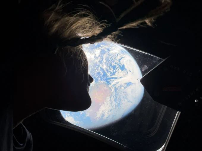 Inside the dim Orion spacecraft, a window in the center of the image shows a brightly lit Earth and an astronaut sits on the right side looking back at Earth. Credit: NASA