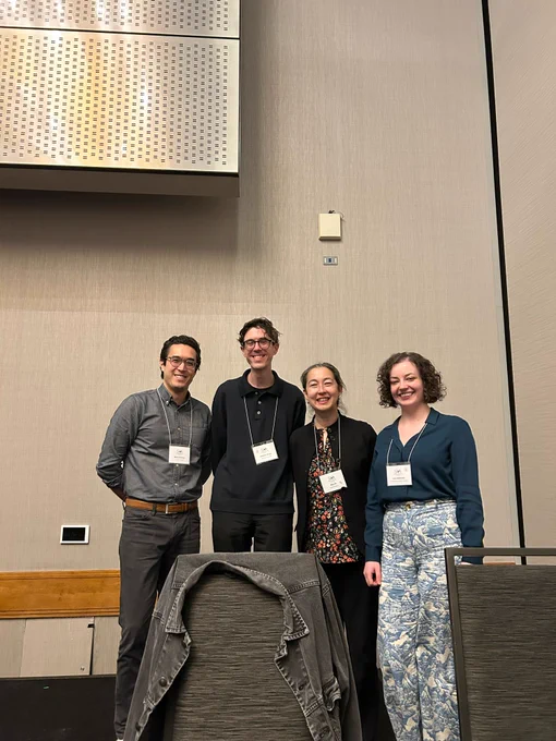 Image of four academics standing behind a table and chair. End ID.