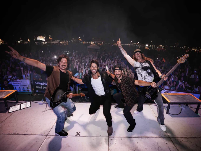 The band Parmalee performing energetically on an outdoor stage at night, with a large crowd in the background. The members are holding guitars and a microphone, posing playfully. Festival lights can be seen behind the audience, creating a lively atmosphere.